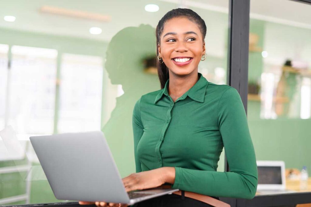 Purposeful African-American businesswoman in smart casual wear stand in modern coworking space and holding laptop, female freelancer or student with laptop computer indoors and looks away cheerfully